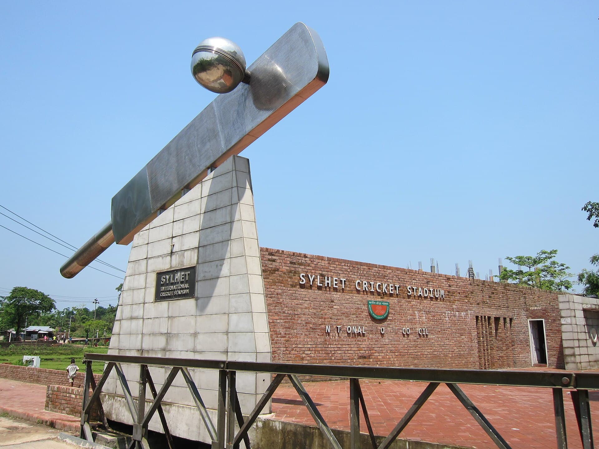 A steel bat and ball sculpture beside the brick wall of Sylhet Cricket Stadium