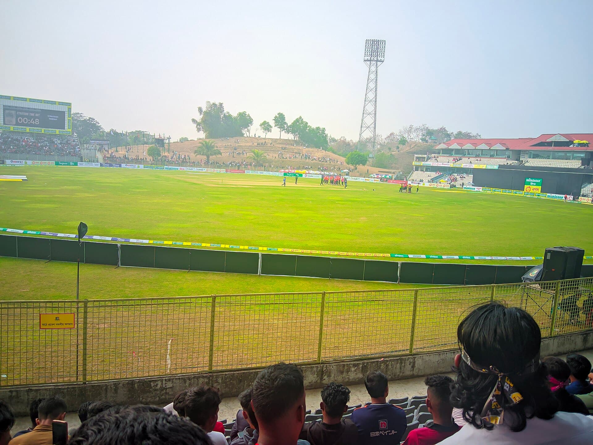 Spectators watching a match, with the grass hill stand and floodlight behind